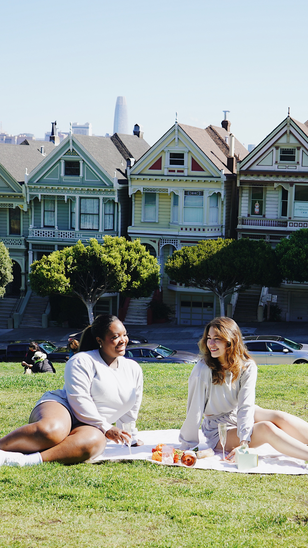A portrait orientation of the two young women, Taysia and Kiara, Asian and Black, are wearing the Fog Grey colored Sweatshorts and Cropped Sweatshirt. They are looking at each other and sitting on the lawn blanket in front of the Painted Ladies. 