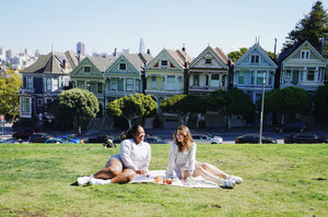 The two young women, Taysia and Kiara, Asian and Black, are wearing the Fog Grey colored Sweatshorts and Cropped Sweatshirt. They are looking and smiling at each other, and sitting on the lawn blanket in front of the Painted Ladies. 
