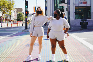 Taysia and Kiara, two young women wearing black masks and standing on the rainbow crosswalk. Taysia leans her arm on Kiara’s shoulder and Kiara looking over her. They are wearing Fog Grey colored Sweatshorts and Cropped Sweatshirt.