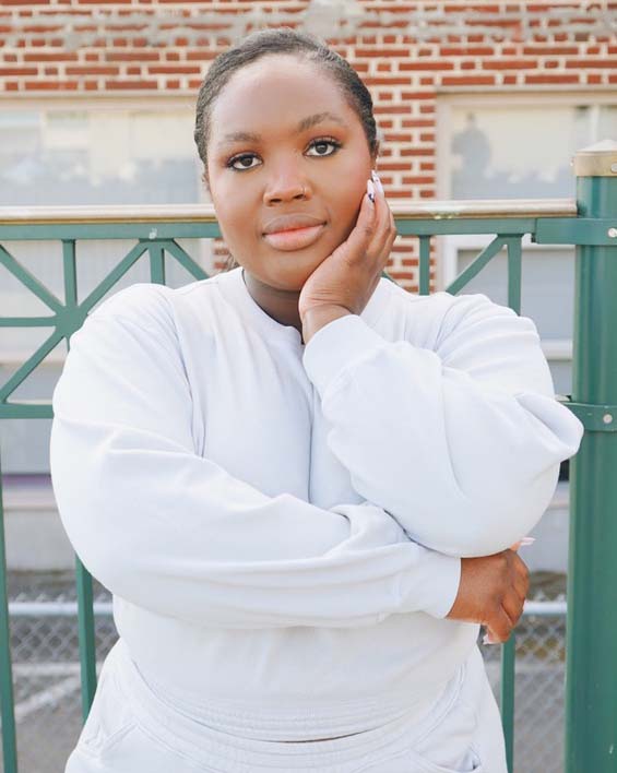 Kiara, a young Black woman wearing the Fog Grey colored Cropped Sweatshirt with red bricks and green fencing in background.