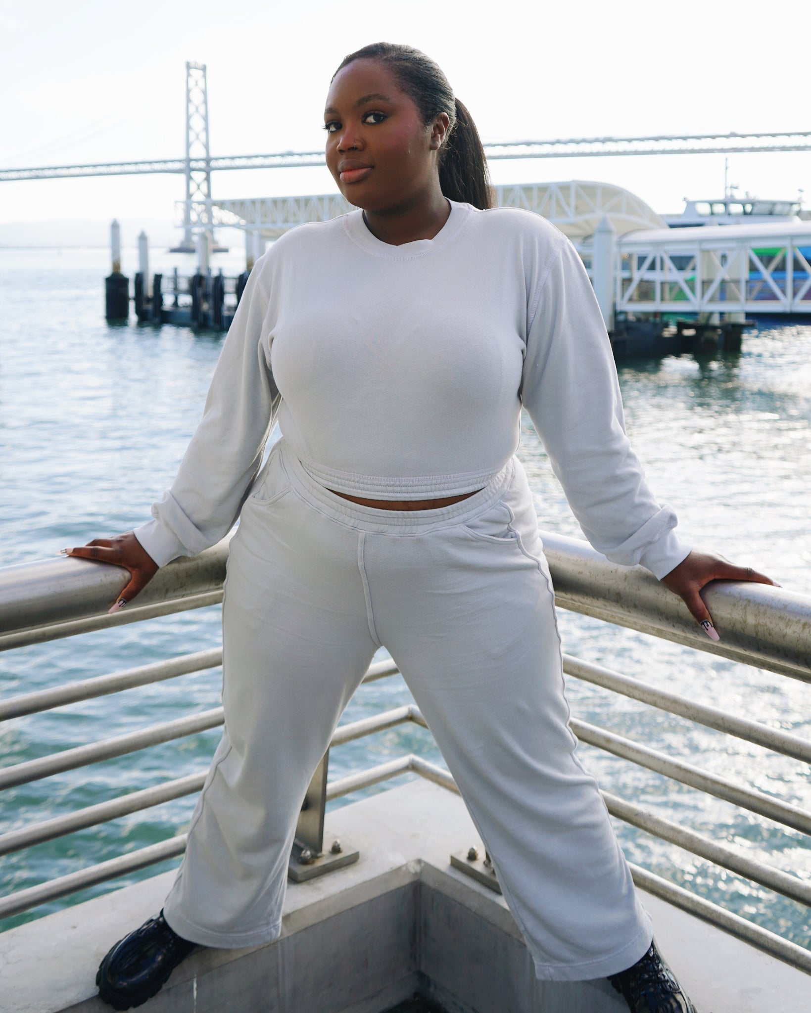 Kiara, a young Black woman is wearing the Fog Grey colored Wide Leg Pants, the Cropped Sweatshirt, and black shoes. She raised herself on a corner of the concrete barrier and is holding onto the metal fence. In the background is the Oakland Bay Bridge, ferry building, and the Bay.