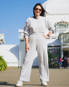Melody, an Asian woman wearing sunglasses showing the Fog Grey colored Wide Leg Pants and Cropped Sweatshirt with white shoes and watch on her right wrist. She is posing in front of the entrance to the Conservatory of Flowers. 
