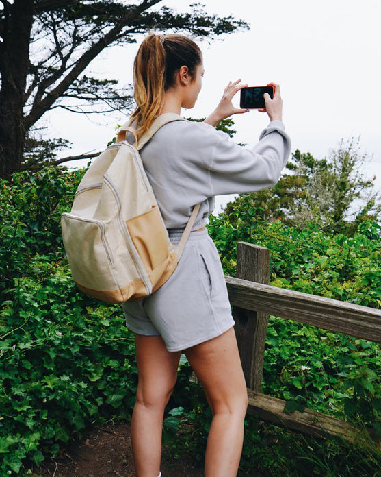 Taysia, a young Asian woman is wearing the Fog Grey colored Sweatshort and Cropped Sweatshirt with tied up dyed brown hair. She has a backpack on her back and standing in front of the bushes, tree, wooden fence and holding a phone to take a picture with her hands. 