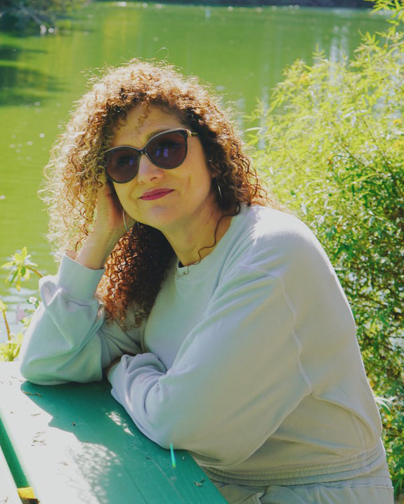 Tiffany, a woman with tan skin and curly hair wearing sunglasses showing the Fog Grey colored Cropped Sweatshirt and sitting on blue picnic table with green lake in background.