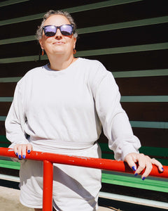 Heidi, a middle-aged Caucasian woman wearing sunglasses and wearing the Fog Grey Cropped Sweatshirt with hands on red metal fencing with green metal horizontal panel in background.