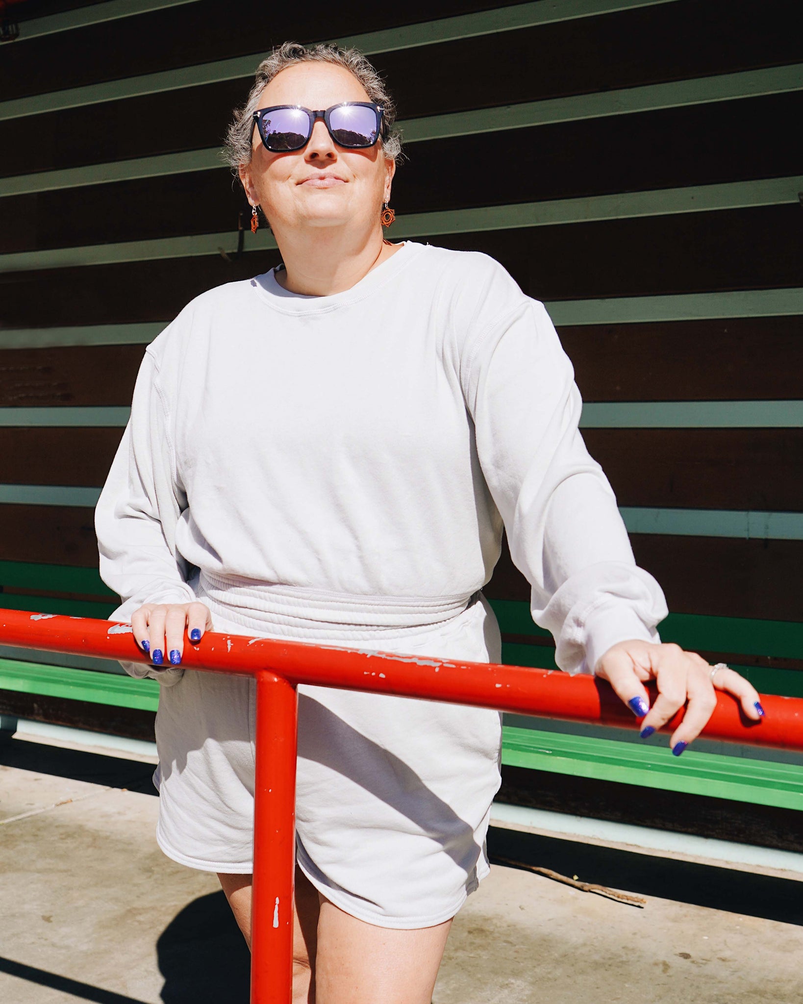 Heidi, a middle-aged Caucasian woman wearing sunglasses and wearing the Fog Grey Sweatshorts along with Cropped Sweatshirt with hands on red metal fencing with green metal horizontal panel in background.