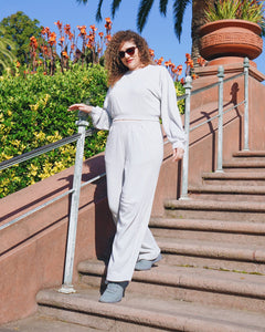 Tiffany, a woman with tan skin and curly hair wearing sunglasses, the Fog Grey colored Wide Leg Pants, the Cropped Sweatshirt, and grey shoes. She is climbing down the stairs in front of the bushes with big pot and palm in the back. 
