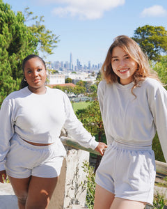The two young women, Taysia and Kiara, Asian and Black, are wearing the Fog Grey colored Sweatshorts and Cropped Sweatshirt. They are standing and leaning against the fences. The location is a park with the city of San Francisco in the background.