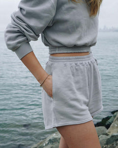 Taysia, a young Asian woman, is wearing the Fog Grey colored Sweatshorts, with gold bracelet, and standing in front of the beach on a foggy day. 
