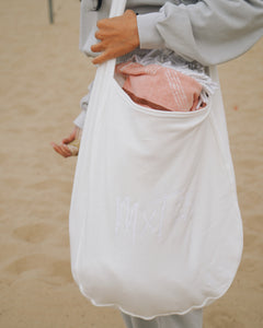 A young woman is wearing the Fog Grey cropped Sweatshirt and pants, holding the oversized tote bag, and standing in the sand. The pink handkerchief is in the tote bag. 